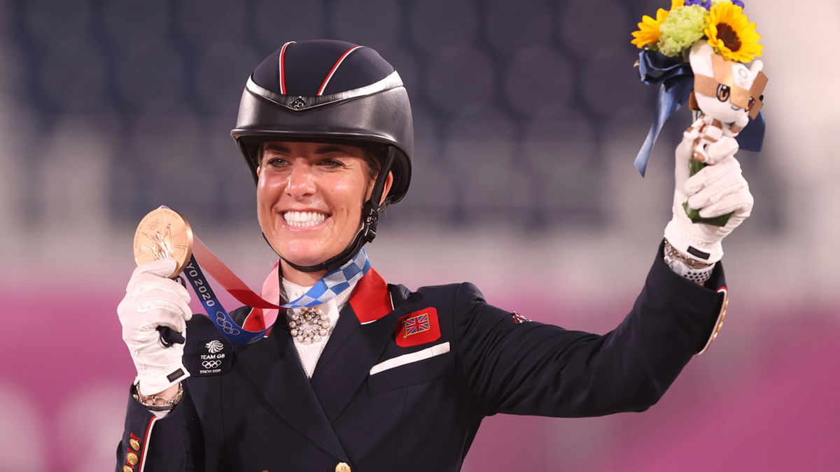 Charlotte Dujardin smiles and holds a bronze medal in her right hand and raises flowers in her left hand