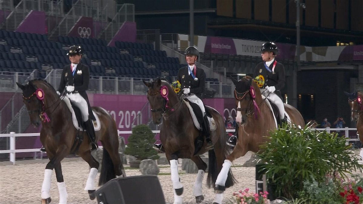 Three members of the German dressage team ride their horses in the arena with ribbons