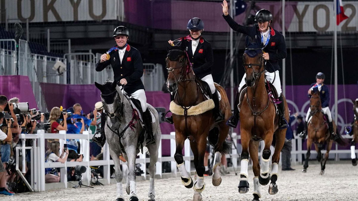 Three people wave and hold medals up as they ride horses