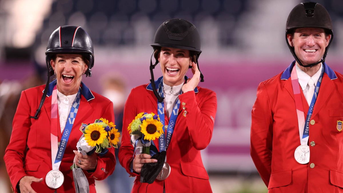 Three riders in red jackets stand smiling and wearing silver medals