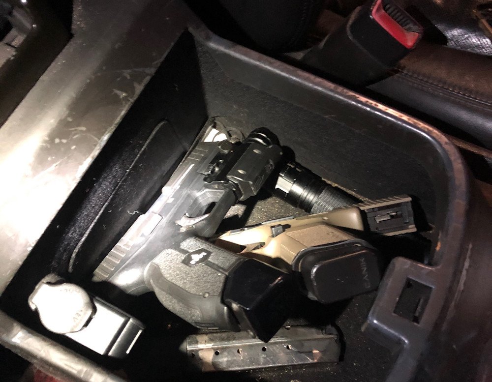 A black handgun over a tan handgun in the center compartment of a vehicle