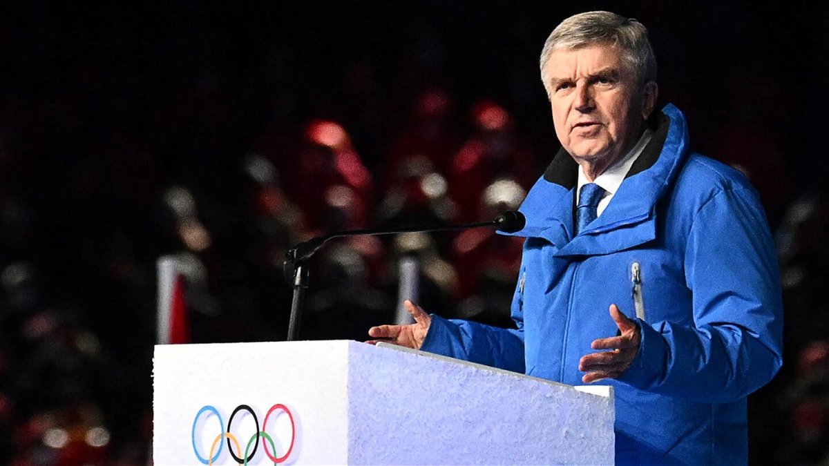 Thomas Bach in blue speaks at white podium with Olympic rings printed on front