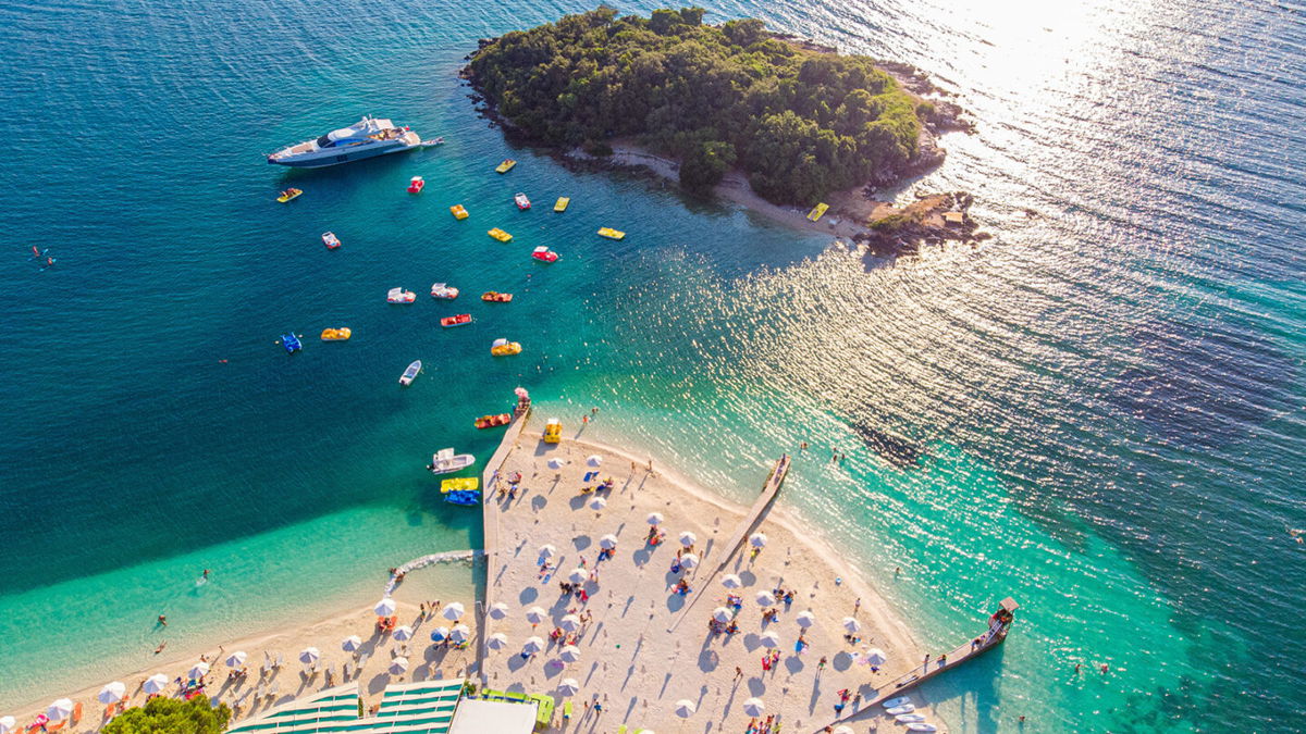 <i>MZaitsev/Adobe Stock</i><br/>Aerial view of a beautiful white sand beach with turquoise water and relaxing people on a sunny day. Ksamil