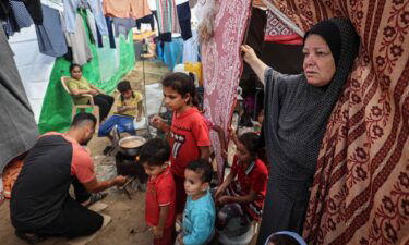 A displaced Palestinian elderly woman stands at the door of her tent with her grandchildren next to her