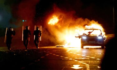 Riot police walks next to a burning police vehicle