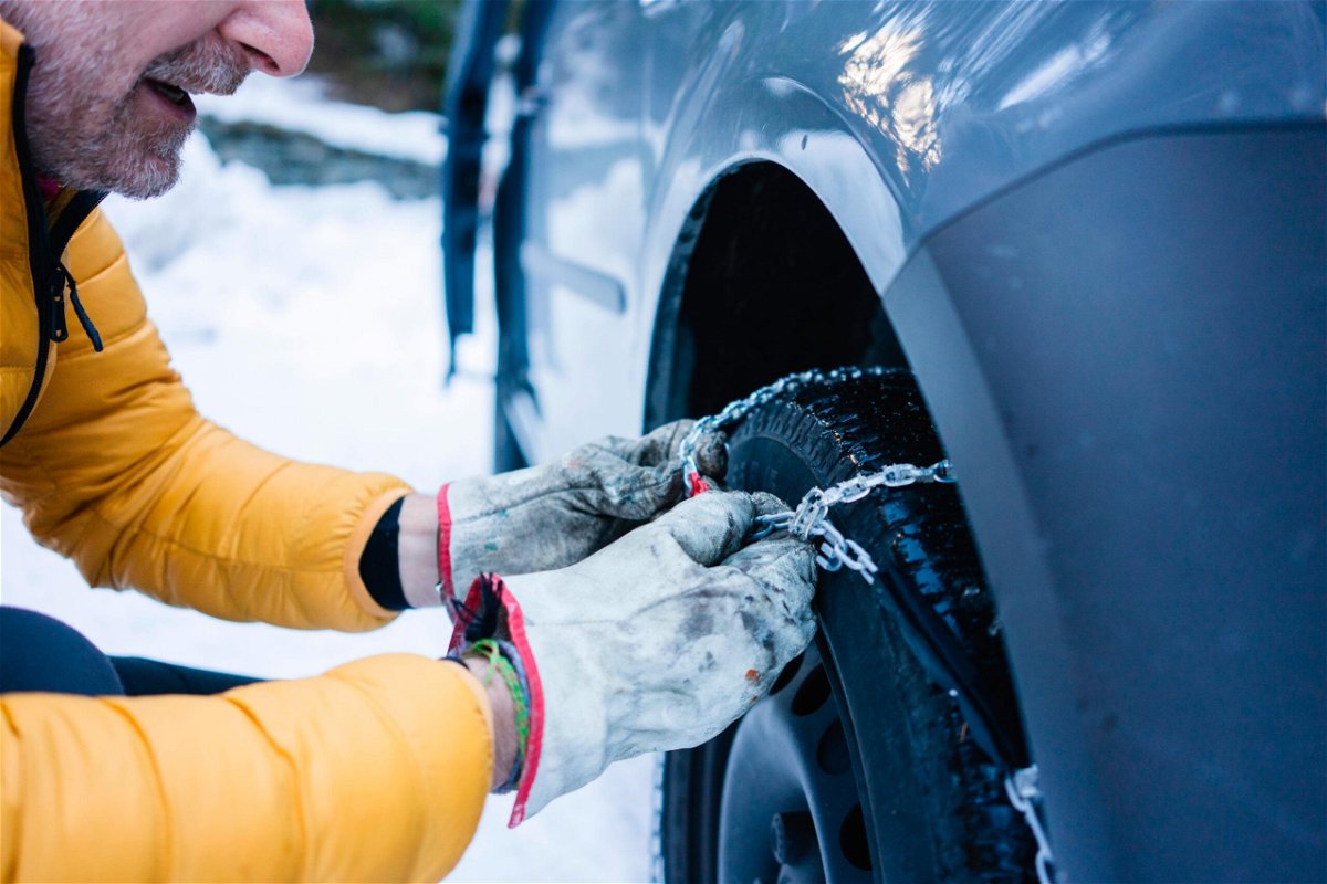<i>Westend61/Getty Images</i><br/>A person puts snow chains on a tire while parked in snowy conditions.