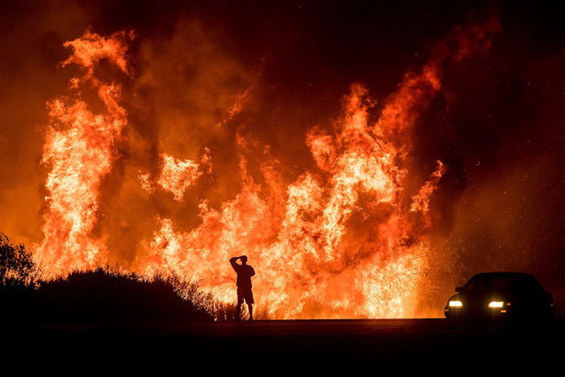 A California utility agrees to pay $80 million over the 2017 Thomas Fire to resolve a federal suit, and here, a motorist watches flames from the Thomas Fire leap above a roadway north of Ventura, California, in December 2017.