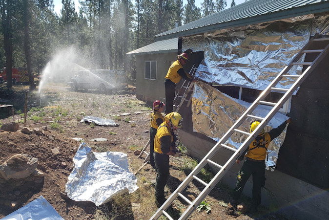 Firefighters from Engine 15, Banks Fire District 13 apply heat-resistant shielding to a home near the western side of the Telephone Fire. They are attached to  TF-47 from the Oregon State Fire Marshal's Structure Protection Group.