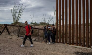 A smuggler stands in the background as asylum-seeking migrants from India cross the Border Wall into the United States from Mexico