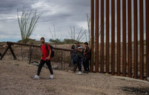 A smuggler stands in the background as asylum-seeking migrants from India cross the Border Wall into the United States from Mexico