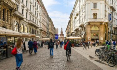 People walking and cycling in the central Via Dante with the entrance tower of the Castello Sforzesco in the background.