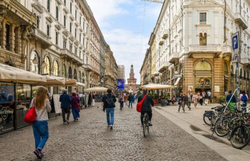 People walking and cycling in the central Via Dante with the entrance tower of the Castello Sforzesco in the background.