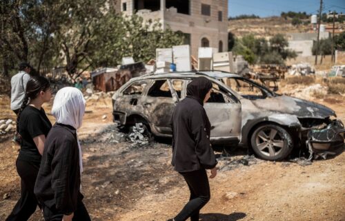 Palestinian women walk past a charred vehicle