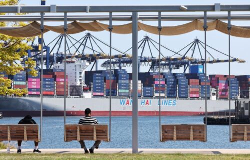 People sit in a park near shipping containers stacked on a container ship at the Port of Los Angeles on June 25